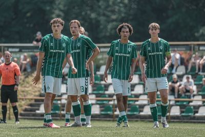 Vier Fußballspieler in grün-weißen Trikots stehen auf dem Spielfeld, im Hintergrund sind ein Schiedsrichter und Zuschauer zu sehen.