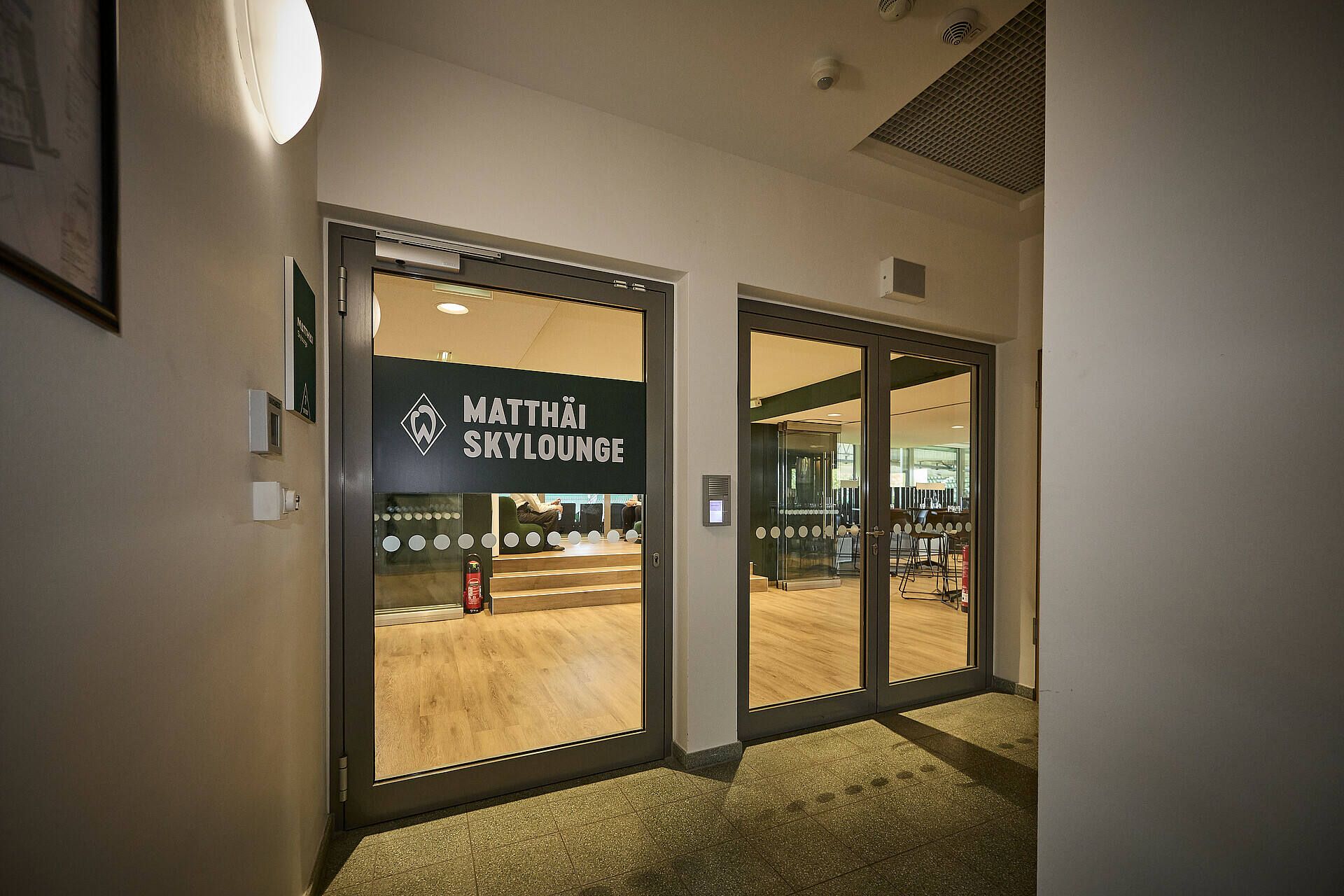 Eingang zur Matthäi Skylounge mit Glastüren, einem Schild darüber und einem warm beleuchteten Innenraum, der durch die Türen hindurch sichtbar ist.