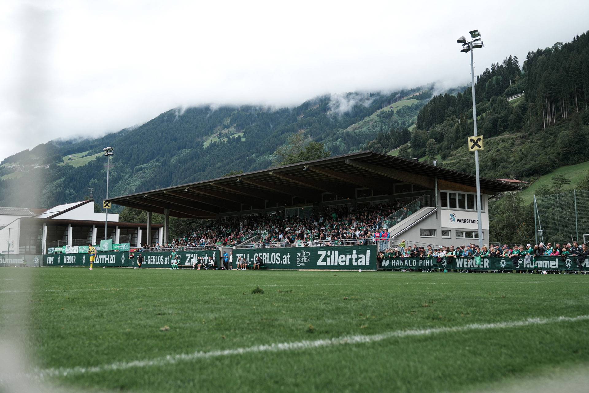 Ein Stadion mit Zuschauern in einer bergigen Landschaft, unter bewölktem Himmel, umgeben von üppig grünen Hügeln und Wäldern.