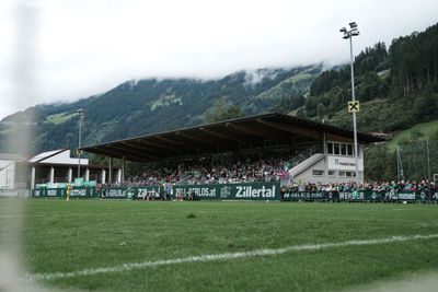 Ein Stadion mit Zuschauern in einer bergigen Landschaft, unter bewölktem Himmel, umgeben von üppig grünen Hügeln und Wäldern.