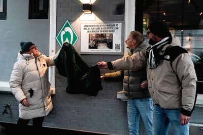 Drei Personen enthüllen ein grünes Tuch, das eine Gedenktafel an einer grauen Backsteinmauer außerhalb eines Gebäudes bedeckt.