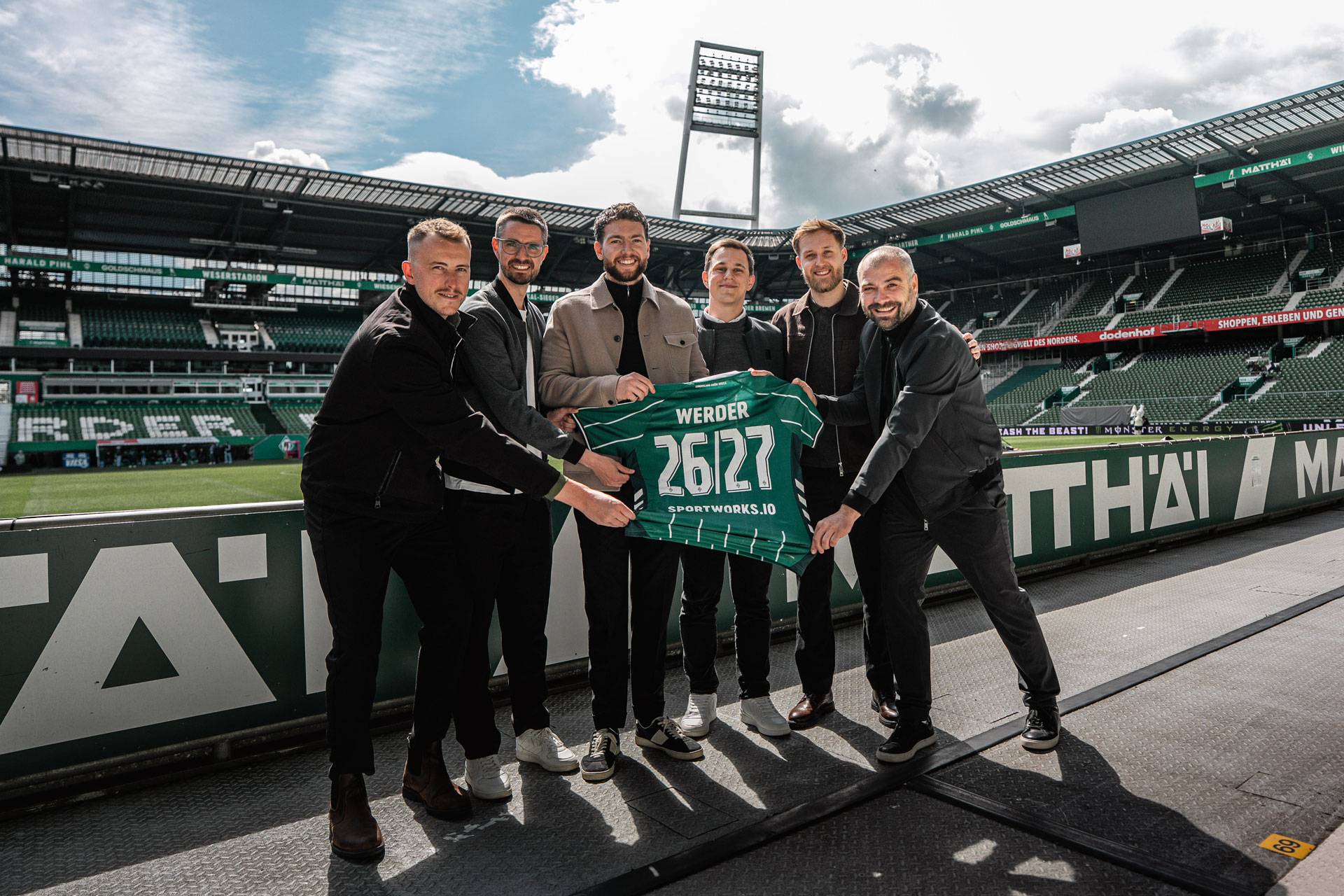 Six people inside the Weserstadion holding up a Werder shirt with 26/27 numbering and sportworks.io printing