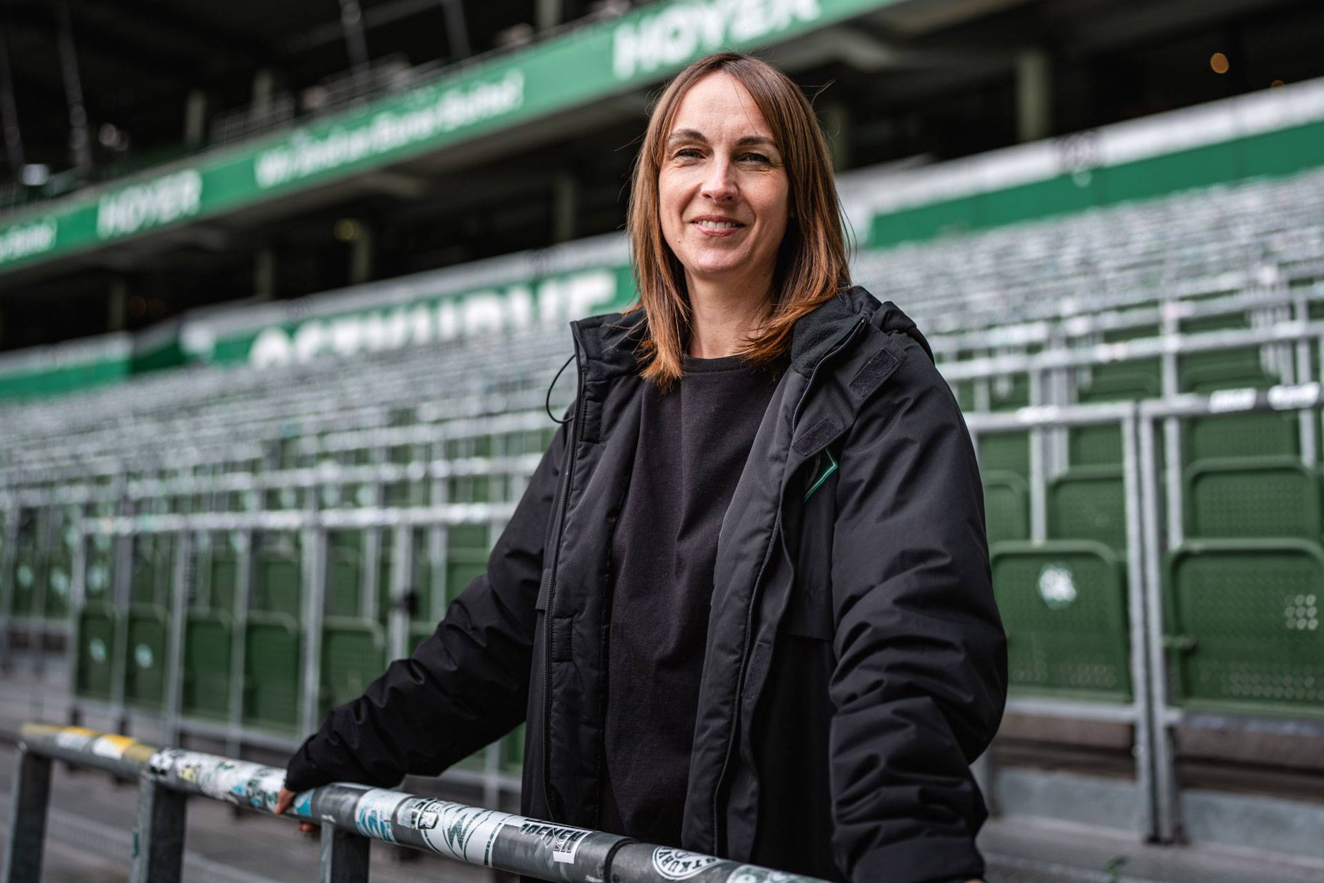 Eine Frau in einer schwarzen Jacke steht lächelnd in einem Stadion voller grüner Sitze.