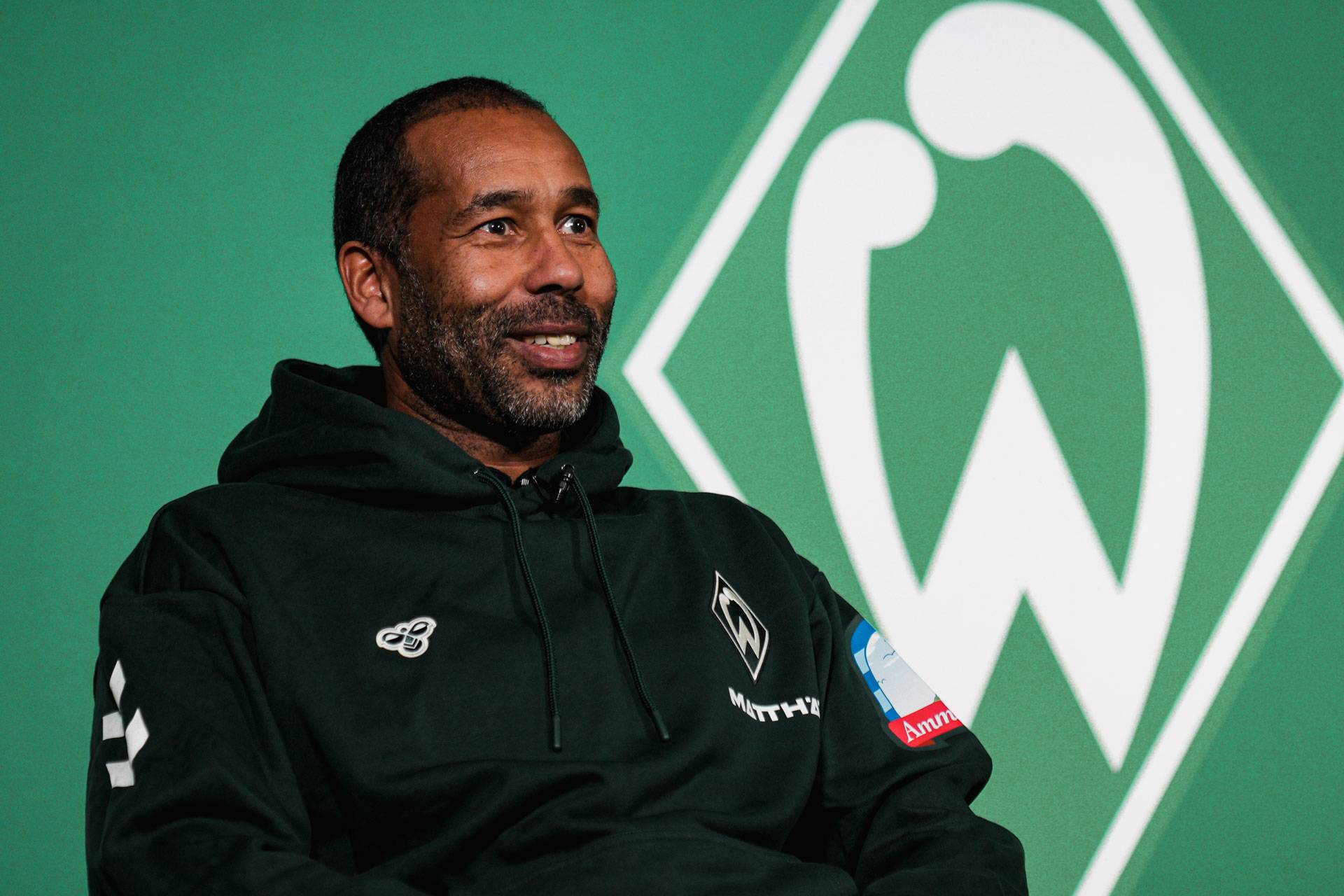 Daniel Thioune in einem dunklen Kapuzenpulli lächelt vor einem grünen Hintergrund mit einem weißen Sportemblem.