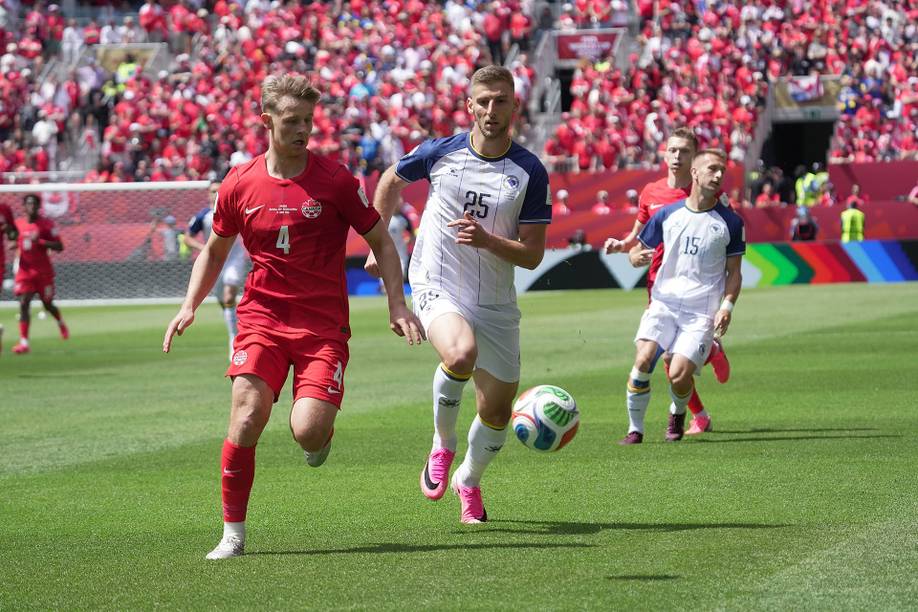 Players in red and white shirts chase the ball on the pitch, with supporters visible in the stands behind them.