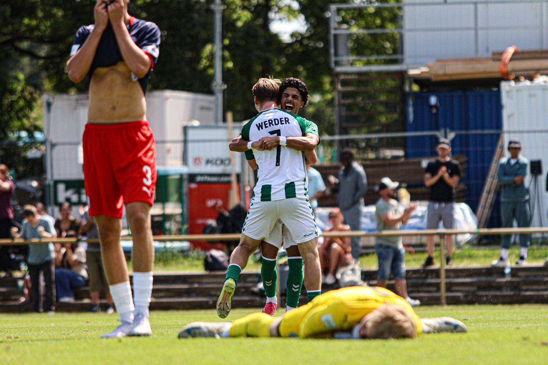 Zwei Fußballspieler in grün-weißen Trikots feiern sich auf dem Spielfeld mit einer Umarmung, während ein niedergeschlagener Spieler in Rot und ein Torwart auf dem Rasen liegen.