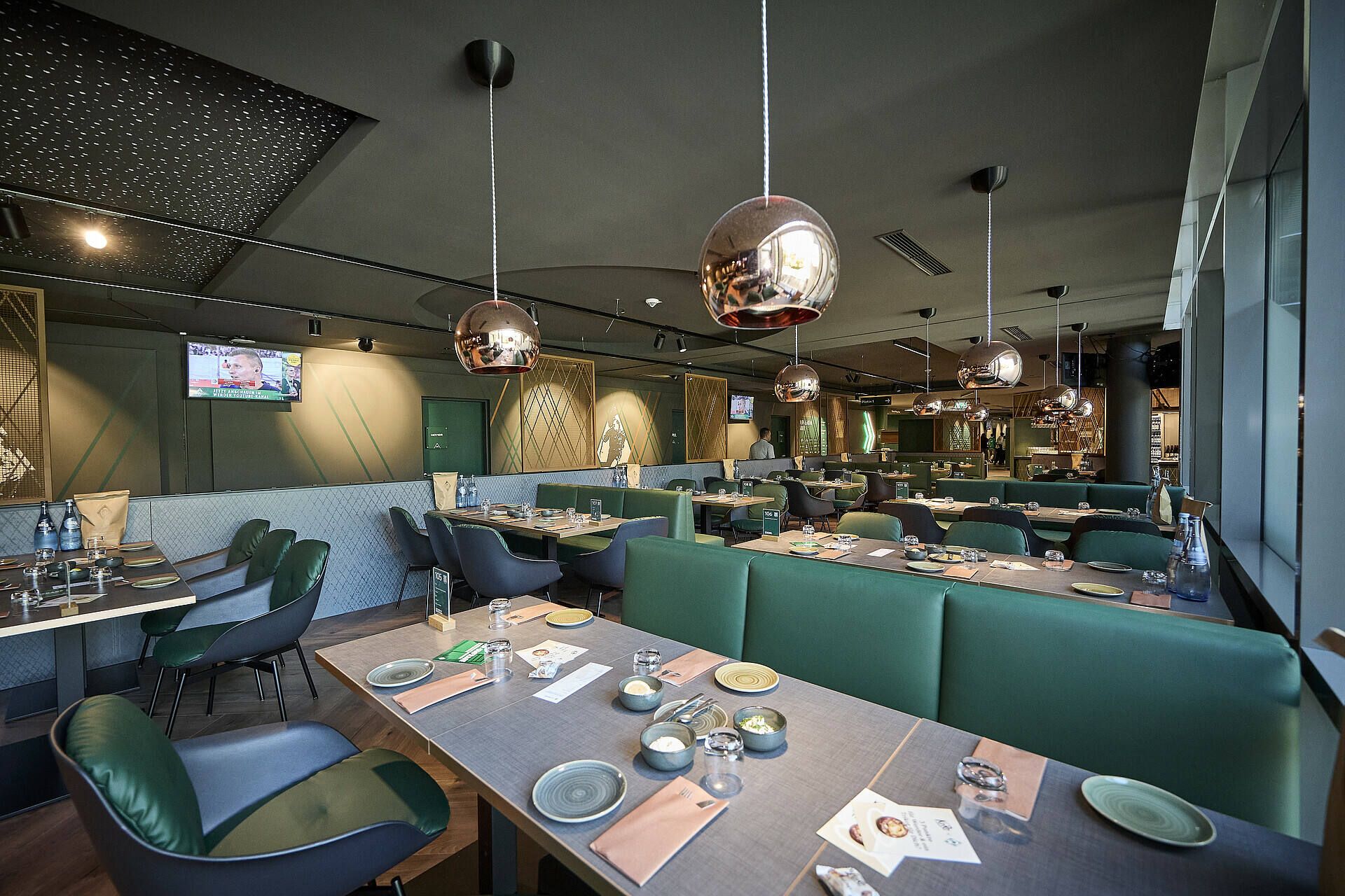 Modernes Restaurantinterieur mit blaugrünen Sitzgelegenheiten, Holztischen und kupferfarbenen Pendelleuchten. Die Tische sind mit Speisekarten, Gläsern und Besteck gedeckt.