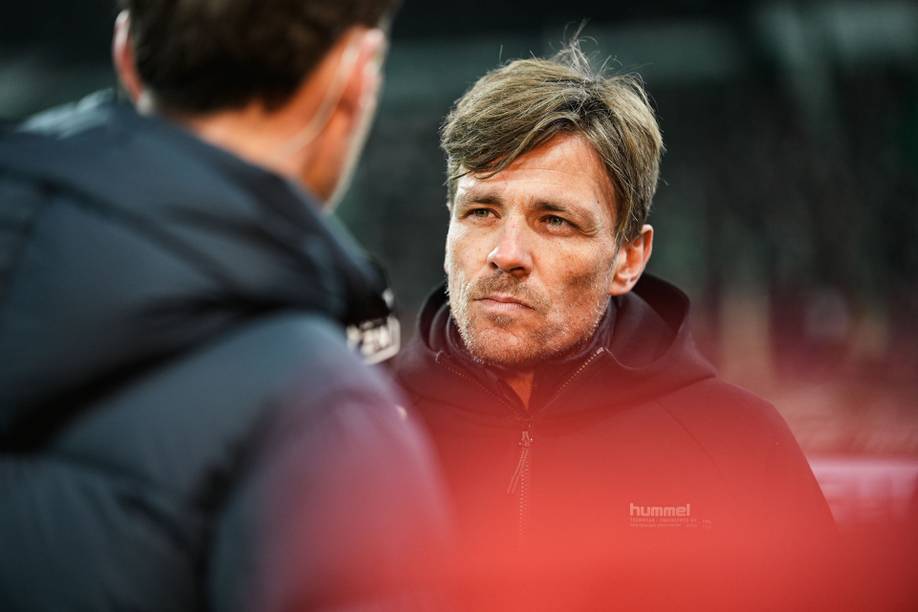 Ein Mann mit kurzen Haaren, der eine schwarze Hummel-Jacke trägt und ernst dreinblickt, während er sich in einer unscharfen Umgebung mit einer anderen Person unterhält.