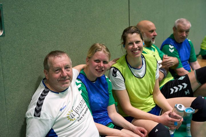 Eine Gruppe von Menschen in Sportbekleidung sitzt an einer Wand, lächelt und ruht sich aus, wobei eine Frau eine Wasserflasche in der Hand hält.