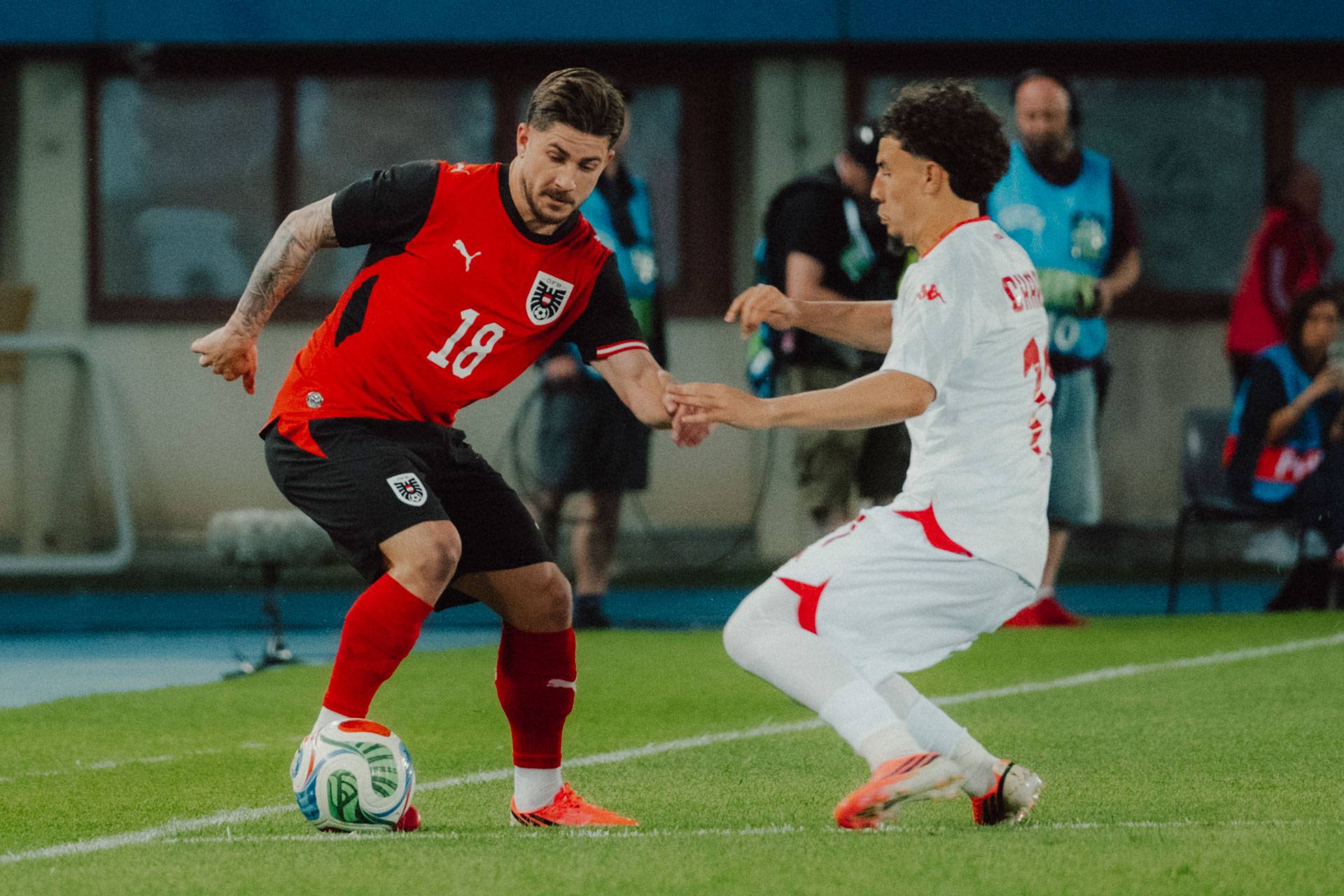 Two footballers in action during a match, one in a red shirt and the other in white, battle for the ball near the touchline.