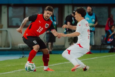 Zwei Fußballspieler in Aktion während eines Spiels, einer im roten Trikot und der andere im weißen, kämpfen nahe der Seitenlinie um den Ball.
