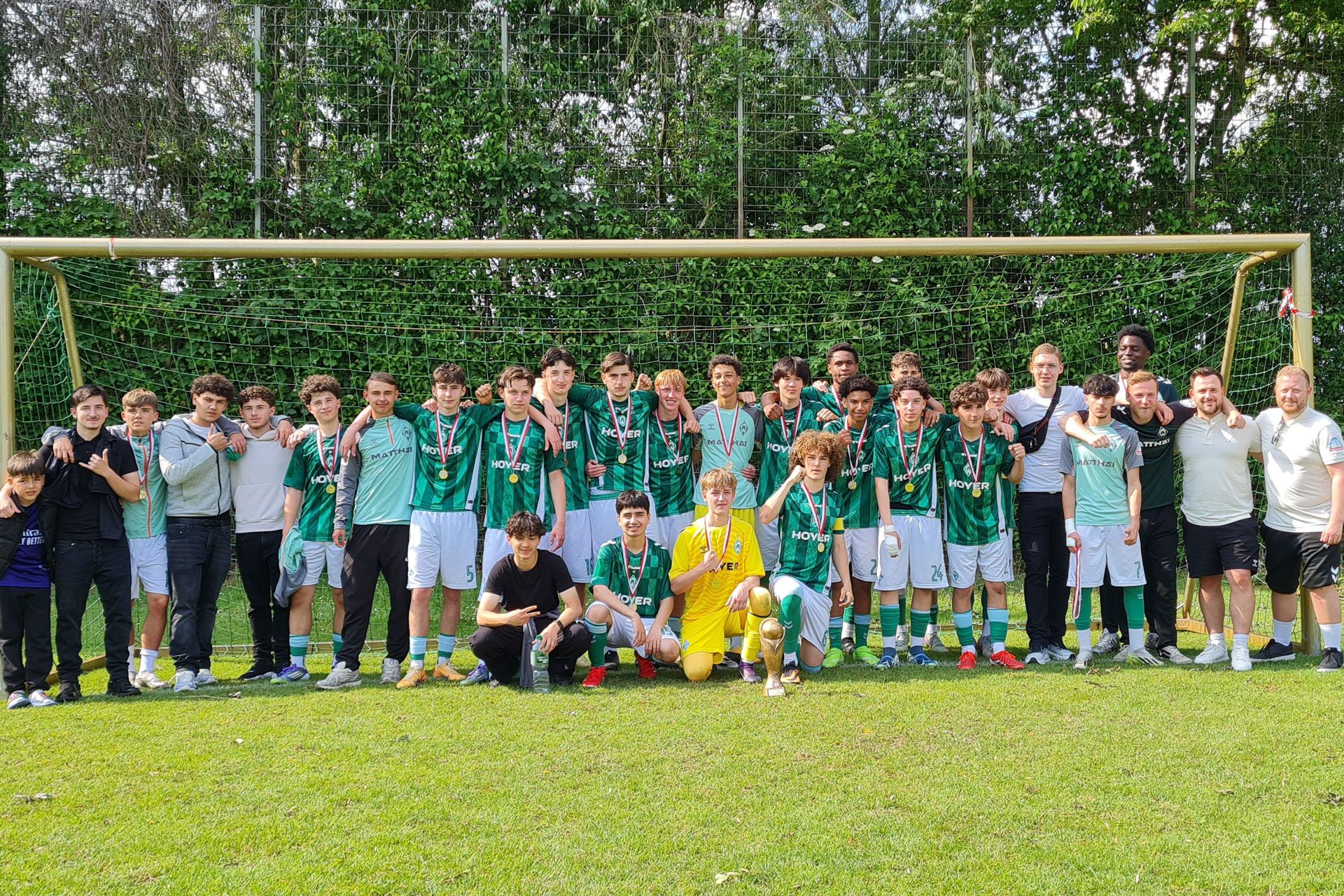 Eine Jugendfußballmannschaft posiert mit Medaillen und einem Pokal vor einem Tor auf einem Rasenplatz, umgeben von Trainern und Betreuern.
