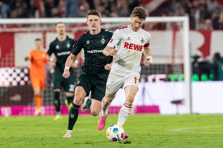 Zwei Fußballspieler in Aktion auf dem Spielfeld, einer in einem weißen Trikot mit der Aufschrift „REWE“ und der andere in einem schwarzen Trikot, im Hintergrund die Zuschauer im Stadion.