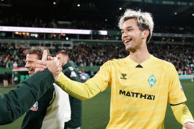 Ein Fußballspieler in einem gelben Trikot lächelt und gibt einem Mann auf dem Spielfeld während eines Spiels die Hand. Im Hintergrund ist die Zuschauermenge zu sehen.