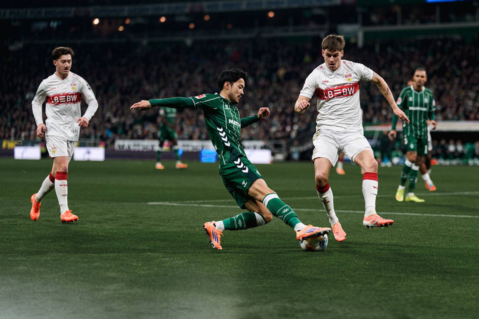 Ein Fußballspieler in grüner Kleidung rutscht auf dem Spielfeld, um einem Spieler in weiß-roter Kleidung den Ball abzunehmen, im Hintergrund sind Zuschauer zu sehen.
