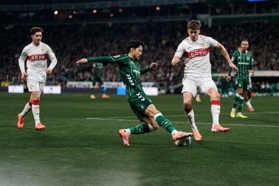 Ein Fußballspieler in grüner Kleidung rutscht auf dem Spielfeld, um einem Spieler in weiß-roter Kleidung den Ball abzunehmen, im Hintergrund sind Zuschauer zu sehen.
