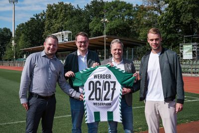 Vier Männer stehen auf einem Fußballplatz und halten ein grün-weißes Fußballtrikot in den Händen, auf dessen Rückseite „Werder 26/27 Weserpark“ aufgedruckt ist.