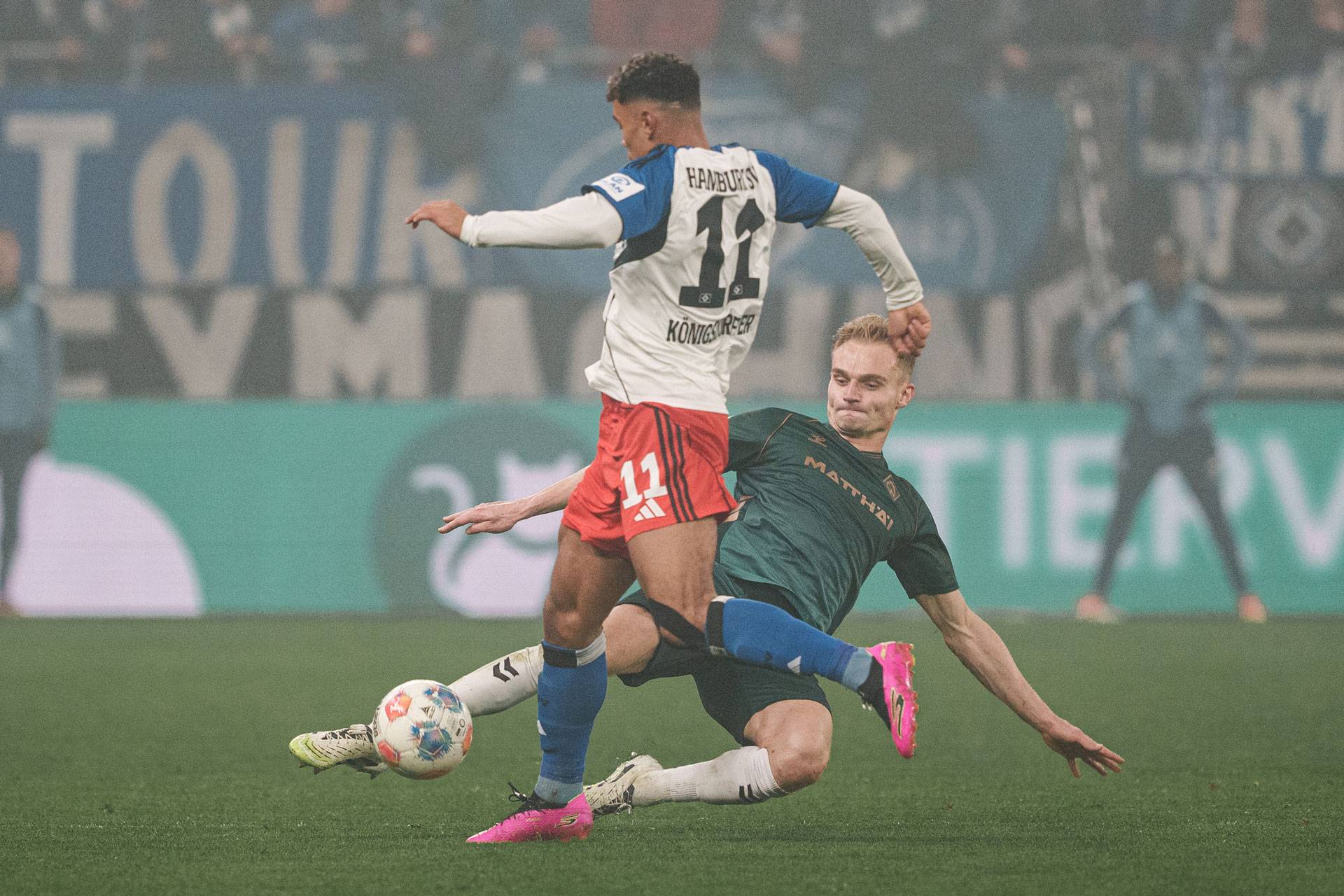 Ein Fußballspieler in roten Shorts fordert während eines Spiels einen Gegner im grünen Trikot heraus, im Hintergrund ist ein vollbesetztes Stadion zu sehen.