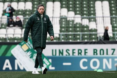Ein Mann in einem grünen Mantel und weißen Schuhen geht über einen Sportplatz, im Hintergrund sind leere Stadionplätze zu sehen.