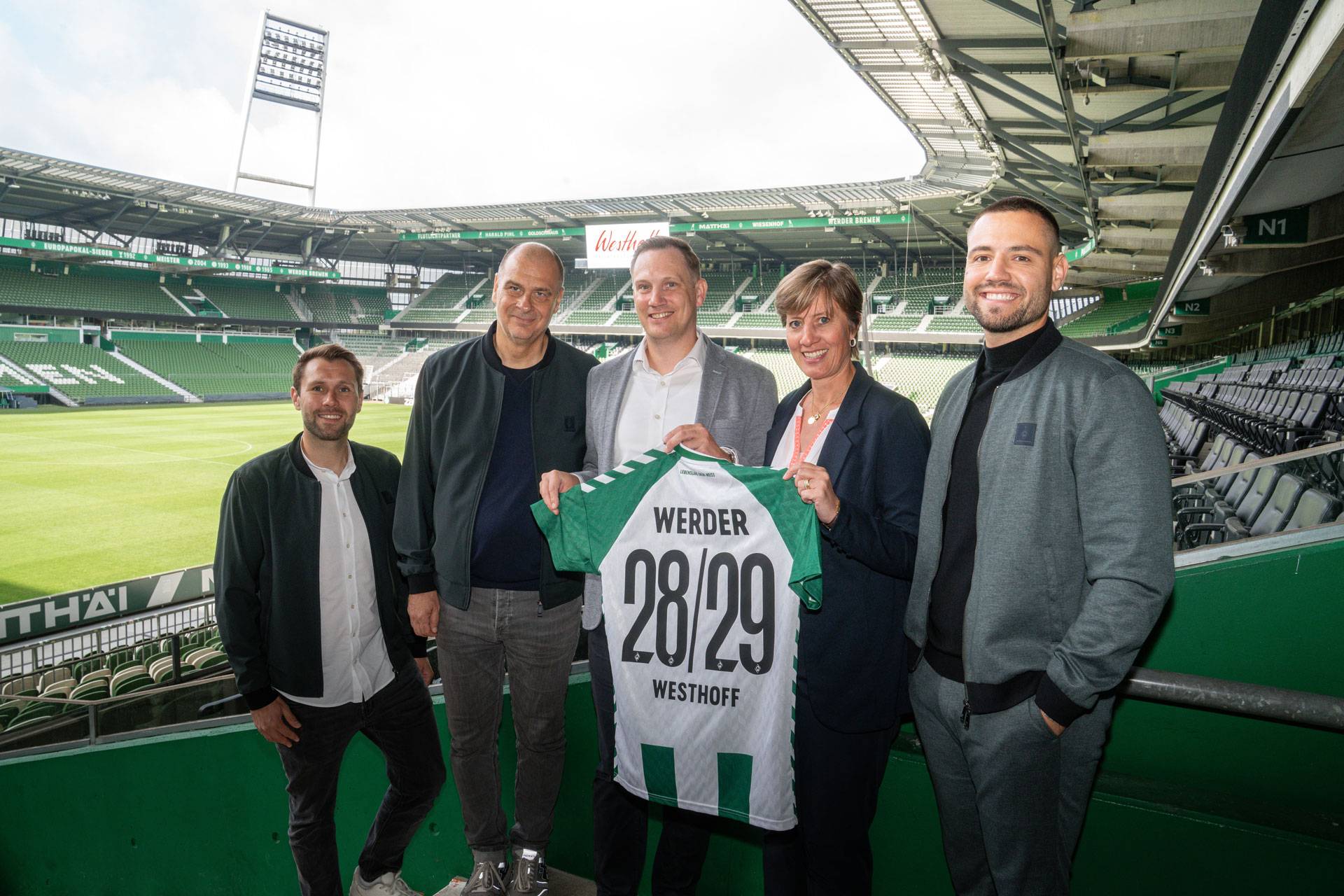 Fünf Personen stehen in einem Stadion und halten ein Trikot von Werder Bremen in den Händen, auf dem die Nummern 28/29 und der Name „Westhoff“ stehen.