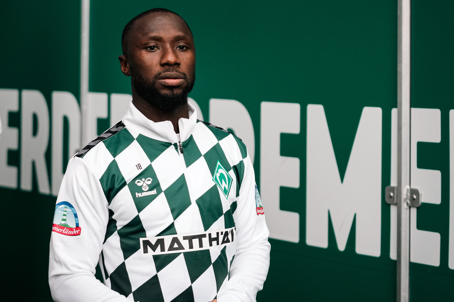Naby Keita steht in einem grün-weiß karierten Trikot vor einem grünen Hintergrund.