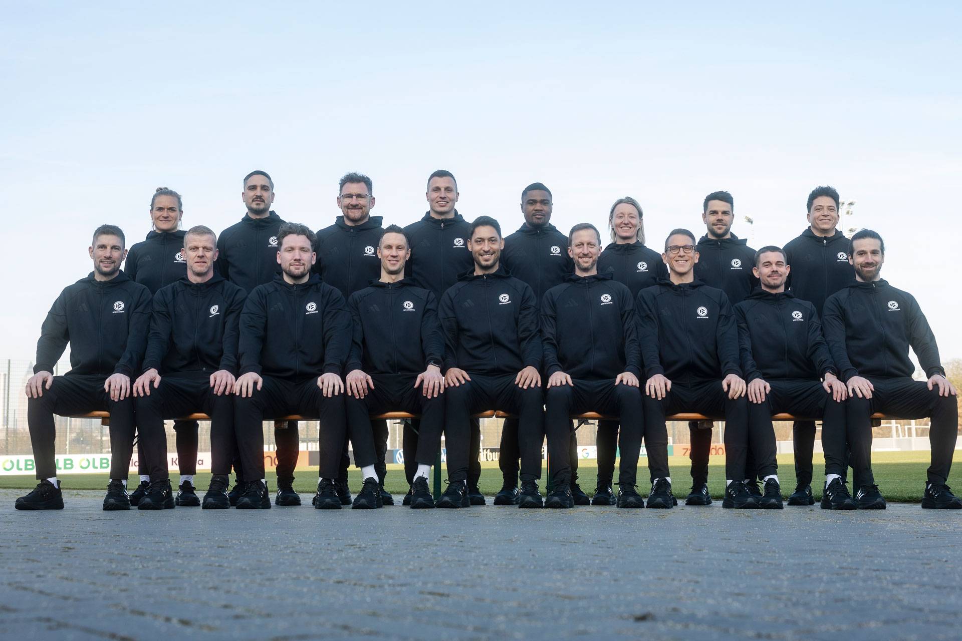 Ein Gruppenfoto von 17 Personen, darunter 3 Frauen, die alle passende schwarze Sportbekleidung tragen und draußen in Reihen sitzen und stehen.