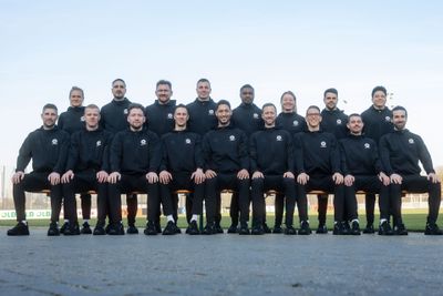 Ein Gruppenfoto von 17 Personen, darunter 3 Frauen, die alle passende schwarze Sportbekleidung tragen und draußen in Reihen sitzen und stehen.