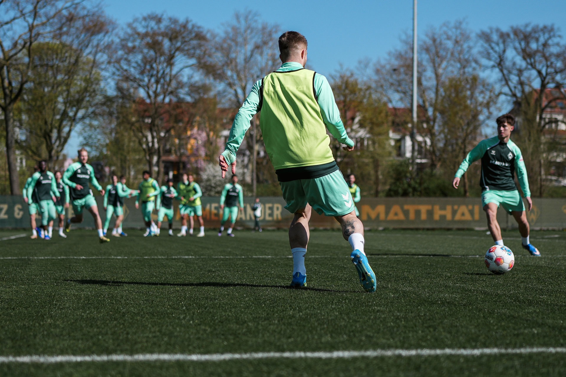 Fußballspieler in grünen Trikots trainieren auf einem sonnigen Platz; einer von ihnen nimmt einen Pass an. Im Hintergrund sind Bäume und Gebäude zu sehen.
