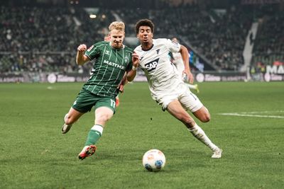 Zwei Fußballspieler, einer in Grün und der andere in Weiß, kämpfen während eines Spiels auf einem Rasenfeld um den Ball.