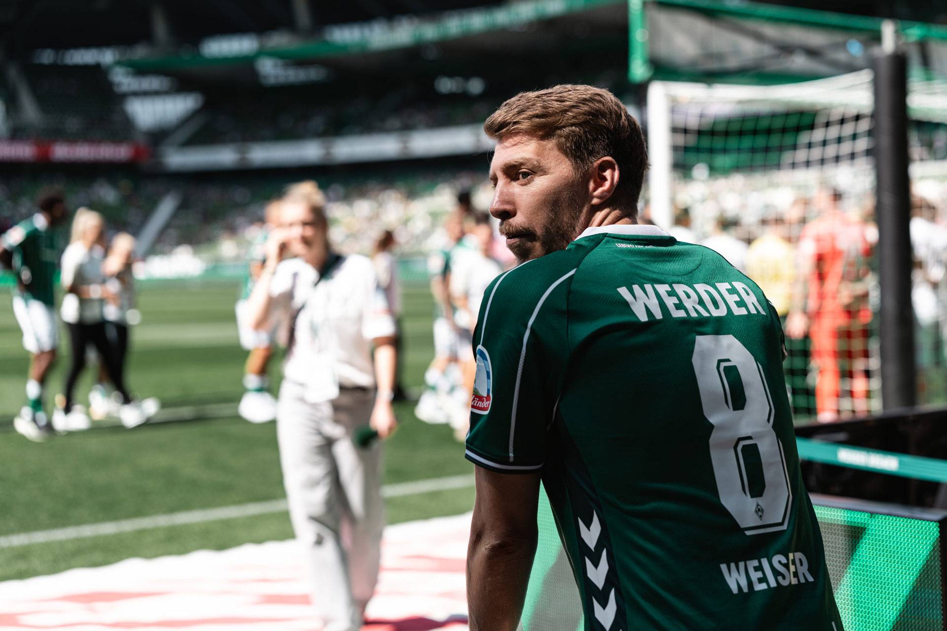 Fußballspieler im grünen Trikot von Werder Bremen mit der Rückennummer 8 auf dem Spielfeld während eines Spiels; im Hintergrund sind verschwommene Menschen und ein Torpfosten zu sehen.