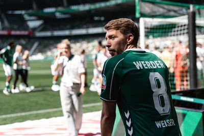 Fußballspieler im grünen Trikot von Werder Bremen mit der Rückennummer 8 auf dem Spielfeld während eines Spiels; im Hintergrund sind verschwommene Menschen und ein Torpfosten zu sehen.