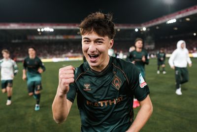 Ein junger Fußballspieler in einem dunkelgrünen Trikot feiert mit erhobener Faust auf dem beleuchteten Stadionrasen, umgeben von seinen Mannschaftskameraden.