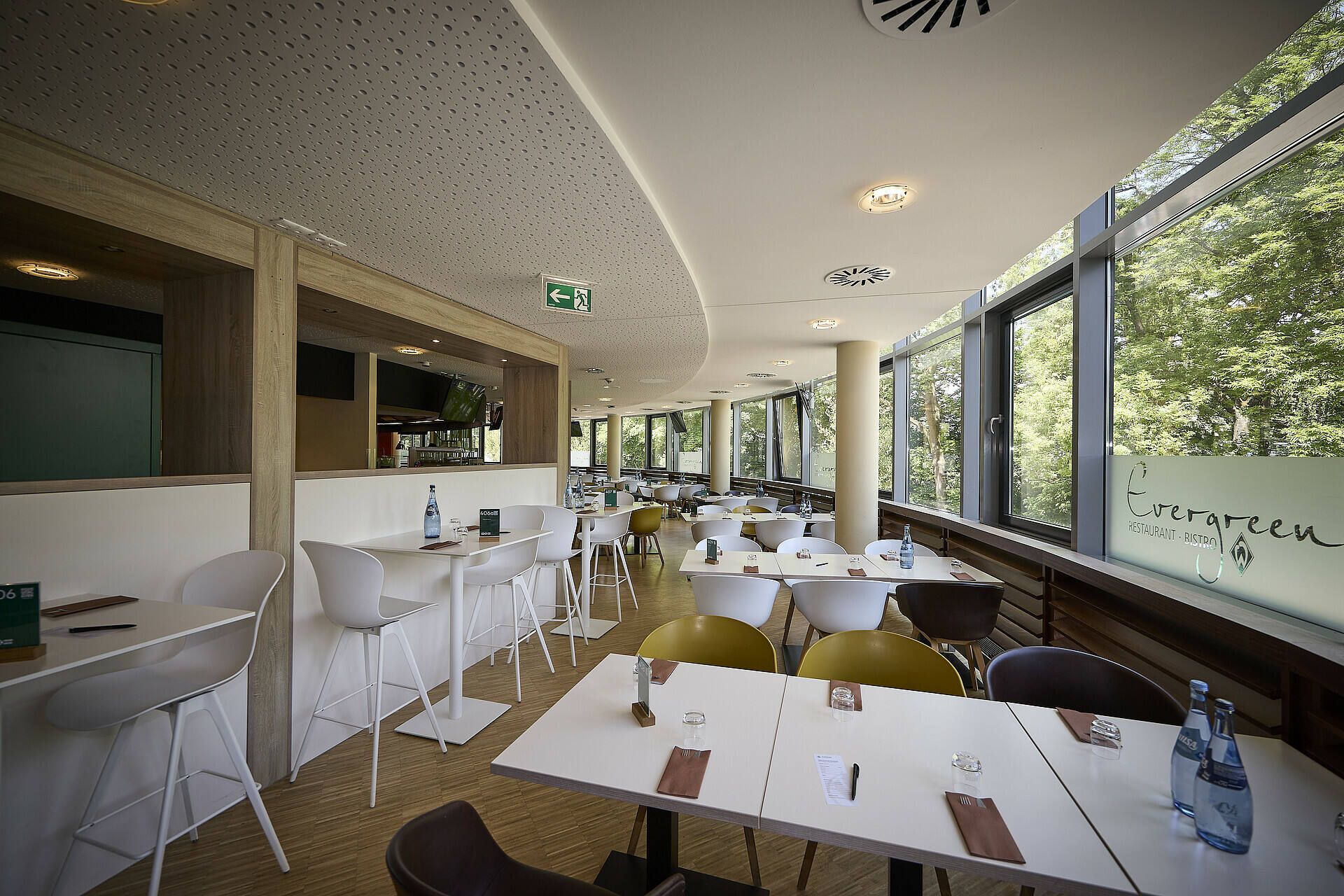 Modernes Café-Interieur mit weißen Tischen, hohen Stühlen und großen Fenstern. Draußen ist Grün zu sehen. Auf den Tischen stehen Flaschen und Besteck.