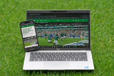 Ein Laptop und ein Smartphone zeigen ein Fußballspiel auf einem Rasenfeld und geben eine Spielszene mit Spielern in Aktion wieder.
