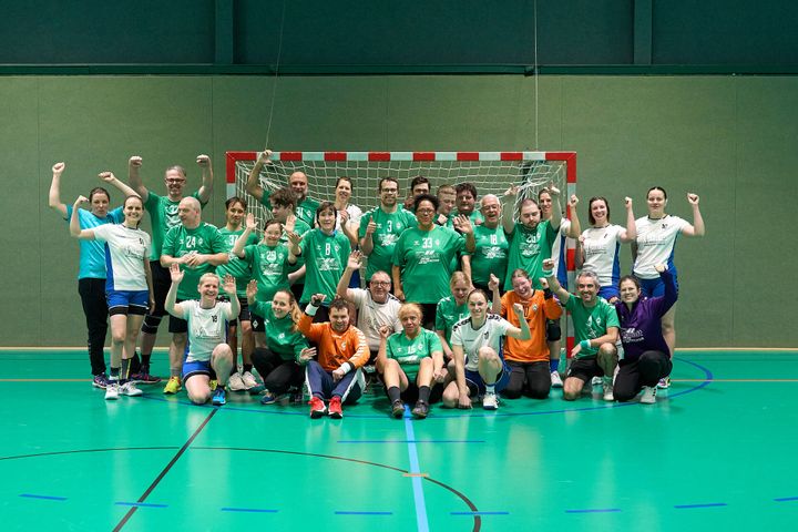Eine Gruppe von Menschen in Sportbekleidung posiert triumphierend in einer Sporthalle, im Hintergrund ist ein Handballtor zu sehen.