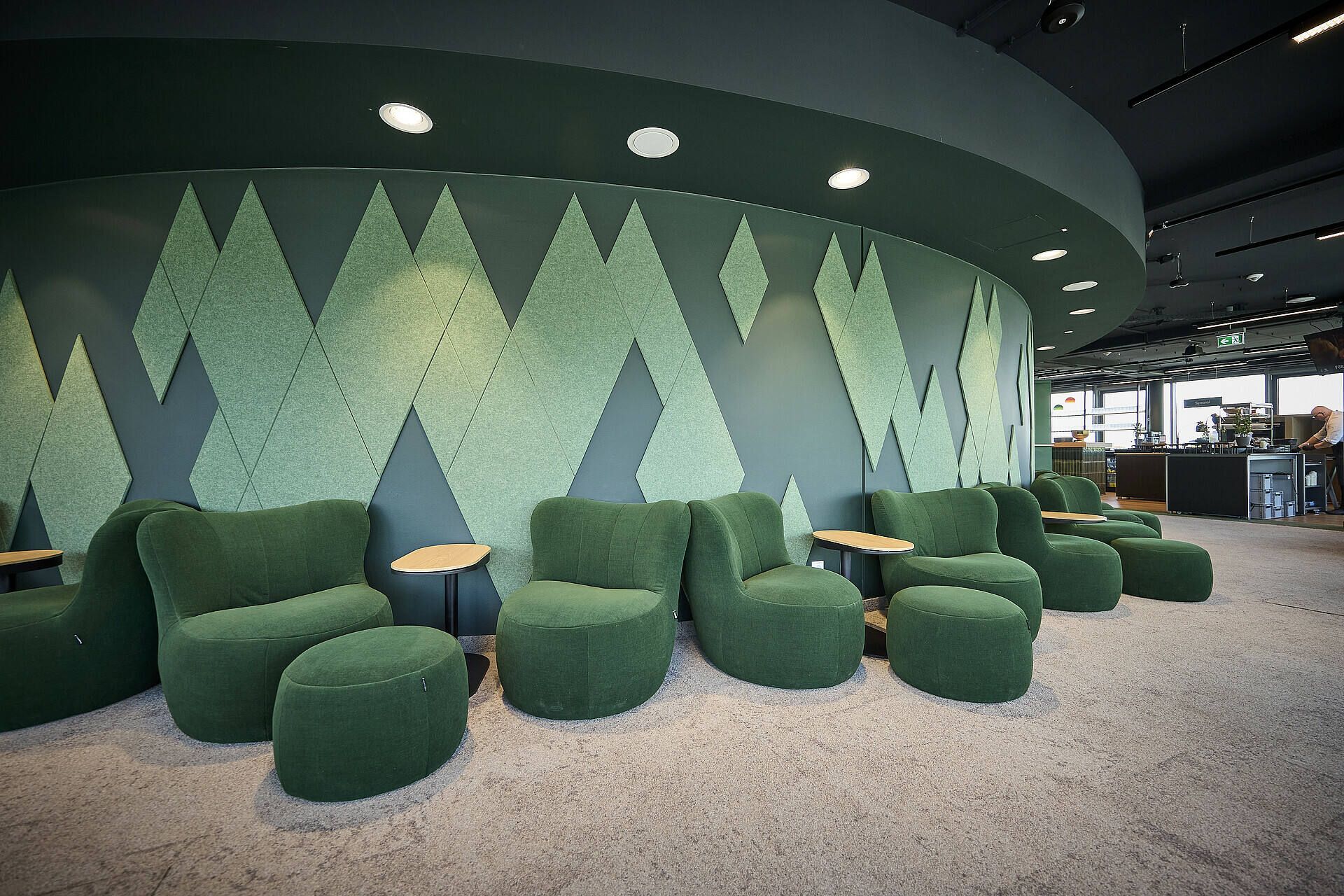 Moderner Loungebereich mit grünen, bequemen Sesseln und geometrischem Wanddesign. Zwischen den Sesseln stehen runde Tische auf einem hellen Teppichboden.