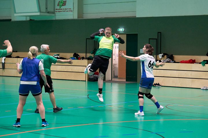 Eine Gruppe von Menschen spielt Hallenhandball; ein Spieler in einem grünen Trikot springt hoch, um den Ball zu werfen, während andere sich auf die Verteidigung vorbereiten.