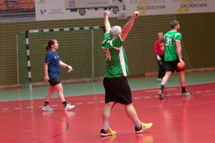 Ein Mann in einem grünen Trikot feiert mit erhobenen Armen auf einem Handballfeld in einer Halle, während andere Spieler im Hintergrund vorbeigehen.