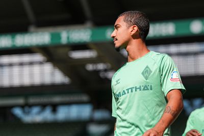 Ein Fußballspieler in einem grünen Trikot mit Sponsorenlogos steht im Freien, blickt in die Ferne, im Hintergrund ist ein verschwommenes Stadion zu sehen.