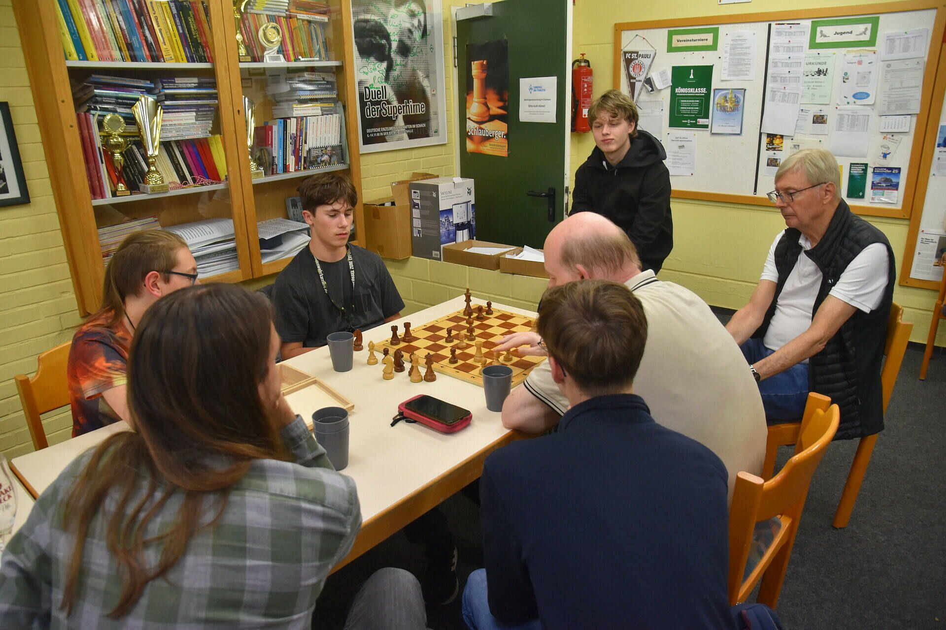 Eine Gruppe von Menschen versammelte sich um ein Schachbrett in einem Raum mit Bücherregalen und Postern und spielte eine Partie.