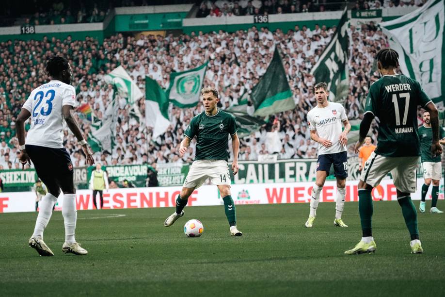 Fußballspiel mit Spielern in Aktion auf dem Spielfeld, umgeben von einer großen Menschenmenge, die grüne und weiße Fahnen schwenkt.