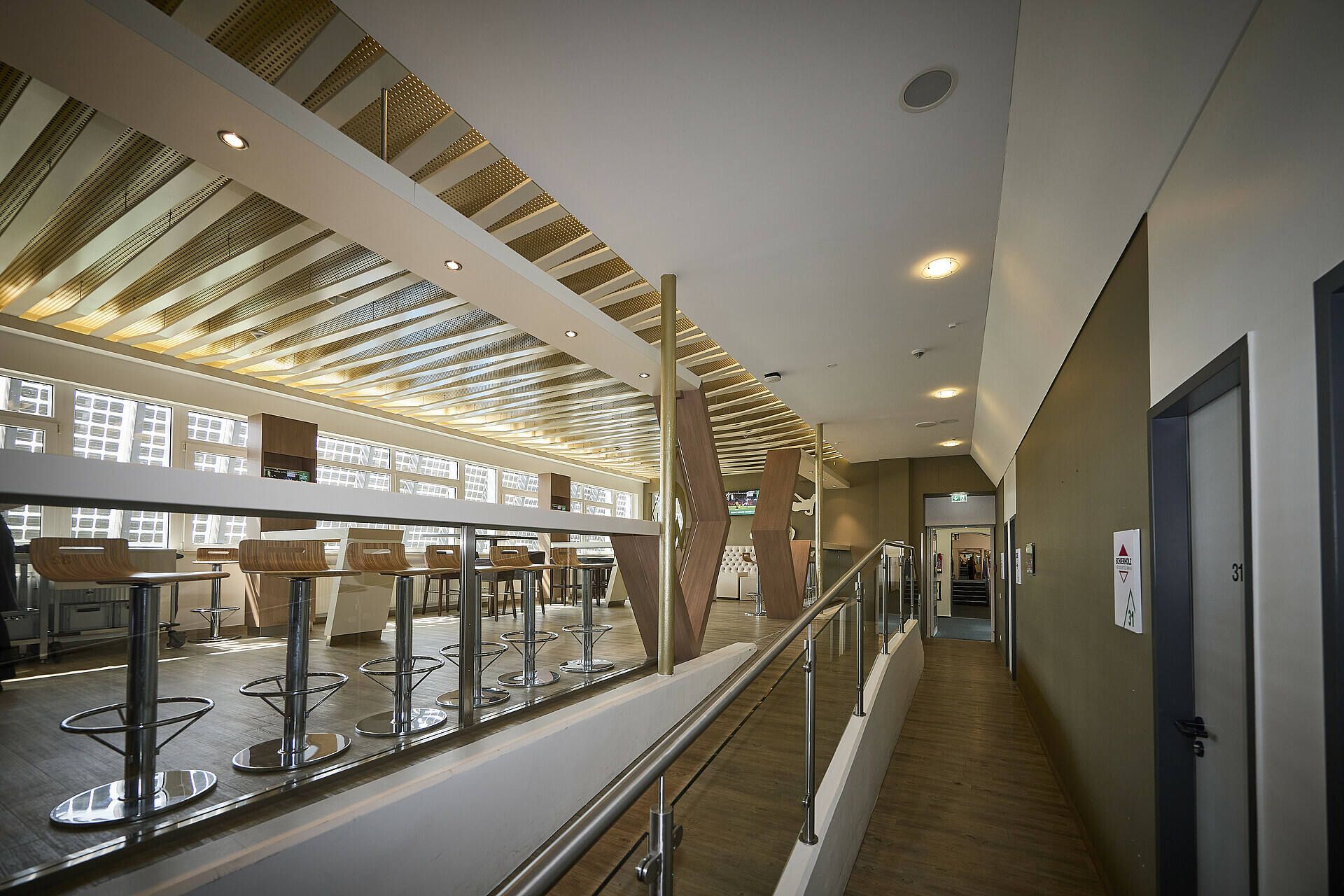 Modernes Interieur mit Holzdecke, Barhockern und einer Rampe, die zu einem Sitzbereich führt. Helle Beleuchtung und große Fenster schaffen eine offene Atmosphäre.