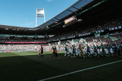 Die Fußballmannschaften betreten das Spielfeld vor einem vollbesetzten Stadion; die Fans schwenken Fahnen, während über ihnen der blaue Himmel strahlt.