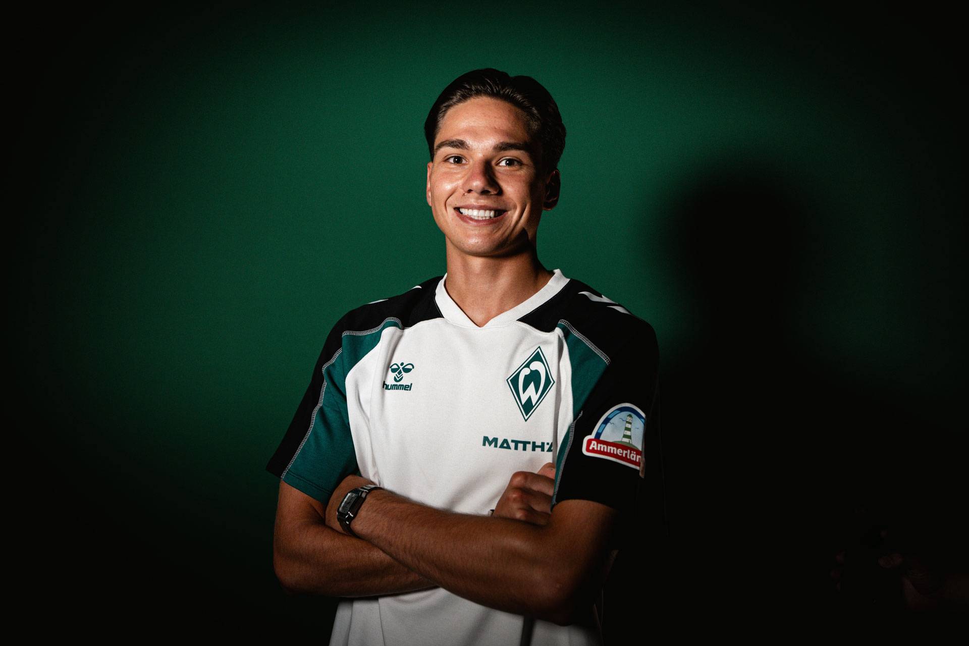 Lächelnder Fußballspieler in einem weiß-grünen Trikot mit verschränkten Armen, der vor einem dunkelgrünen Hintergrund steht.