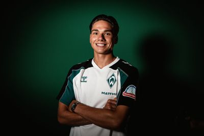 Lächelnder Fußballspieler in einem weiß-grünen Trikot mit verschränkten Armen, der vor einem dunkelgrünen Hintergrund steht.
