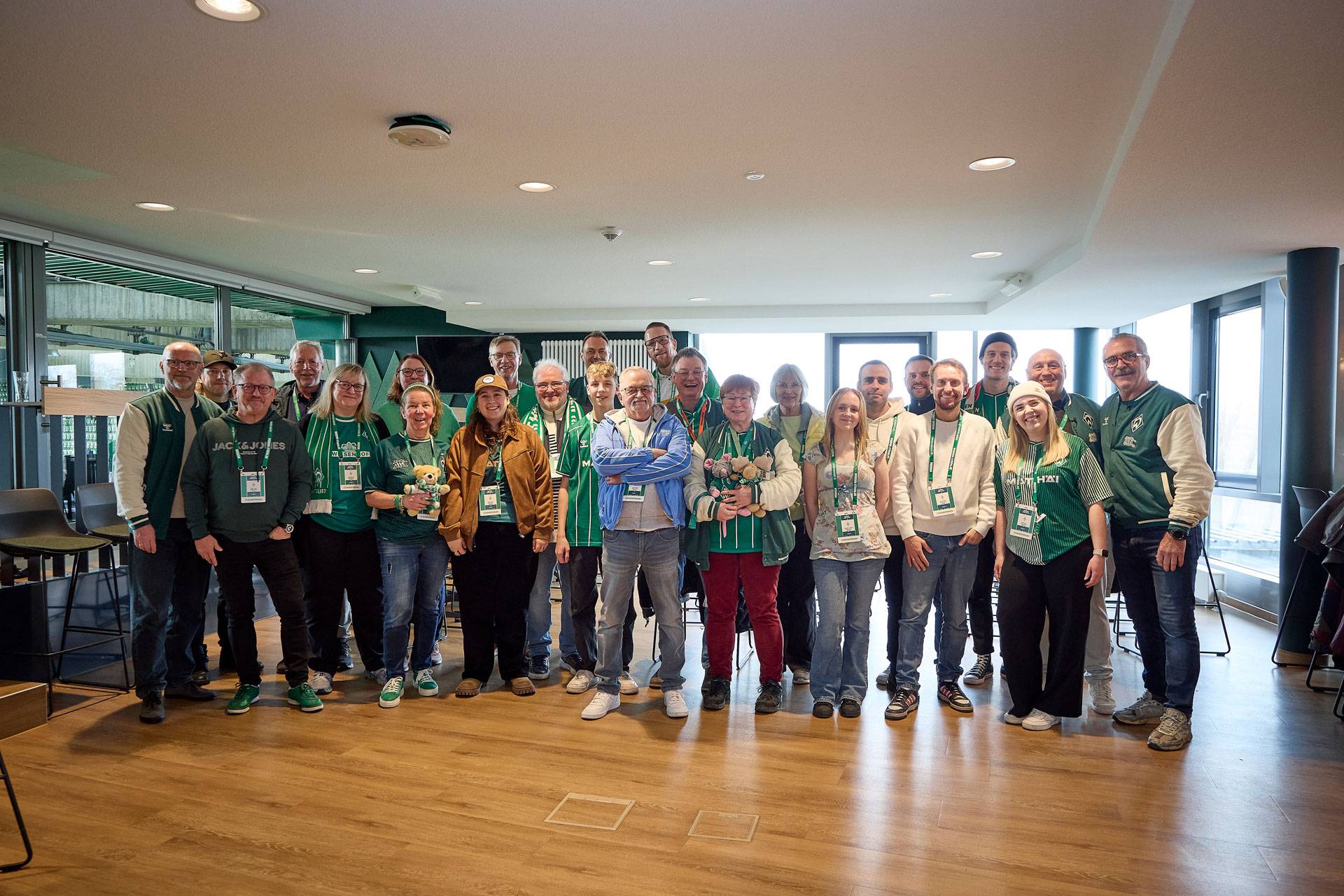 Eine Gruppe von Menschen in grüner Kleidung und mit grünen Ansteckern versammelt sich in einem hell erleuchteten Raum mit Holzboden und lächelt in die Kamera.