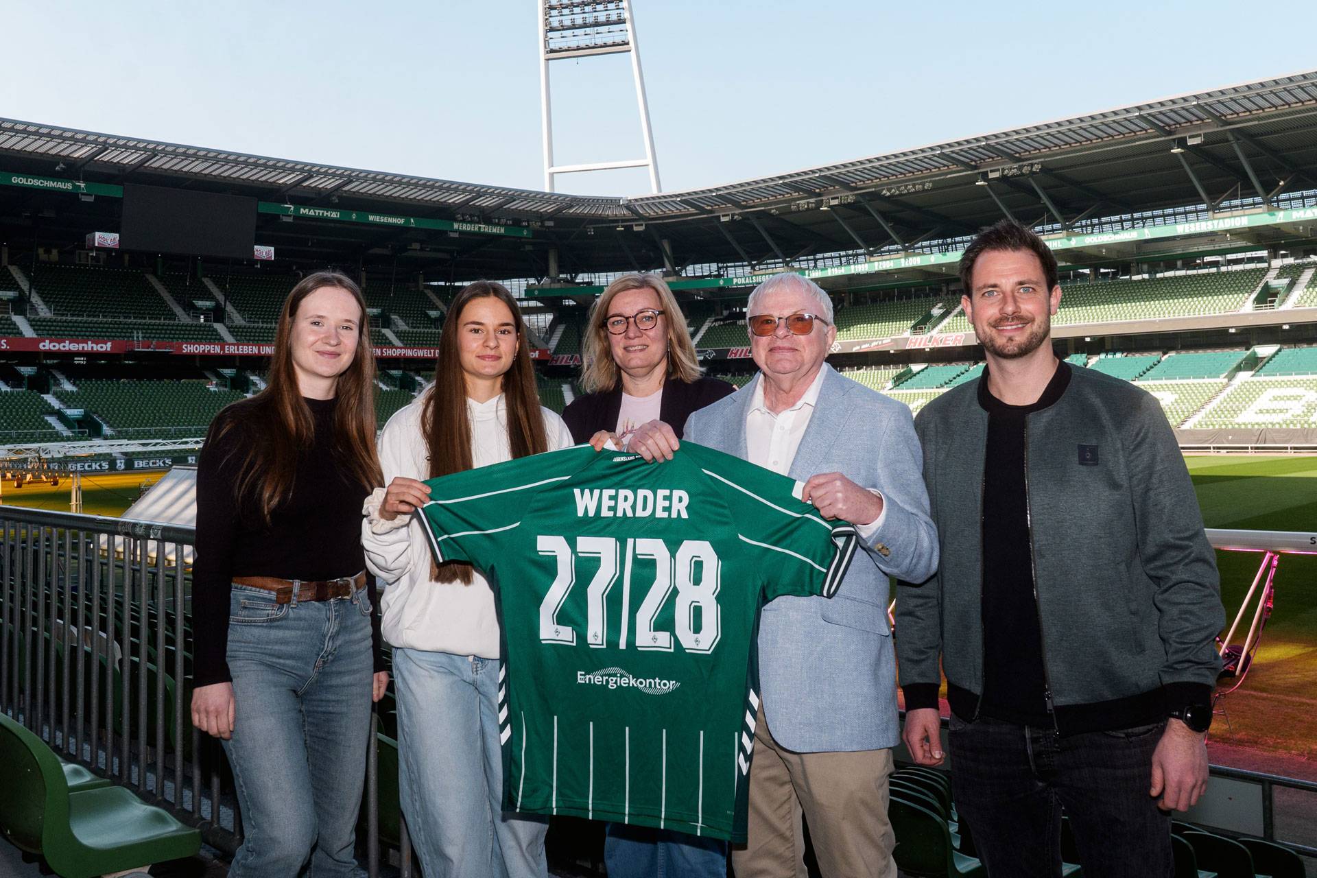 Fünf Personen stehen in einem Stadion und halten ein grünes Fußballtrikot mit der Nummer 27/28 in den Händen.