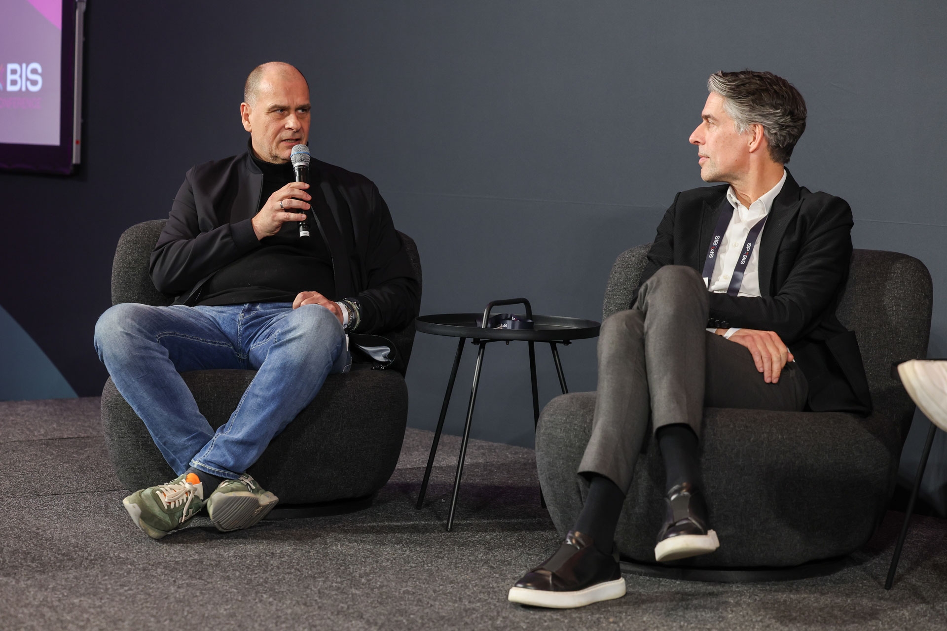 Zwei Männer sitzen auf Bühnenstühlen, einer spricht in ein Mikrofon. Zwischen ihnen steht ein kleiner runder Tisch vor einem dunklen Hintergrund.