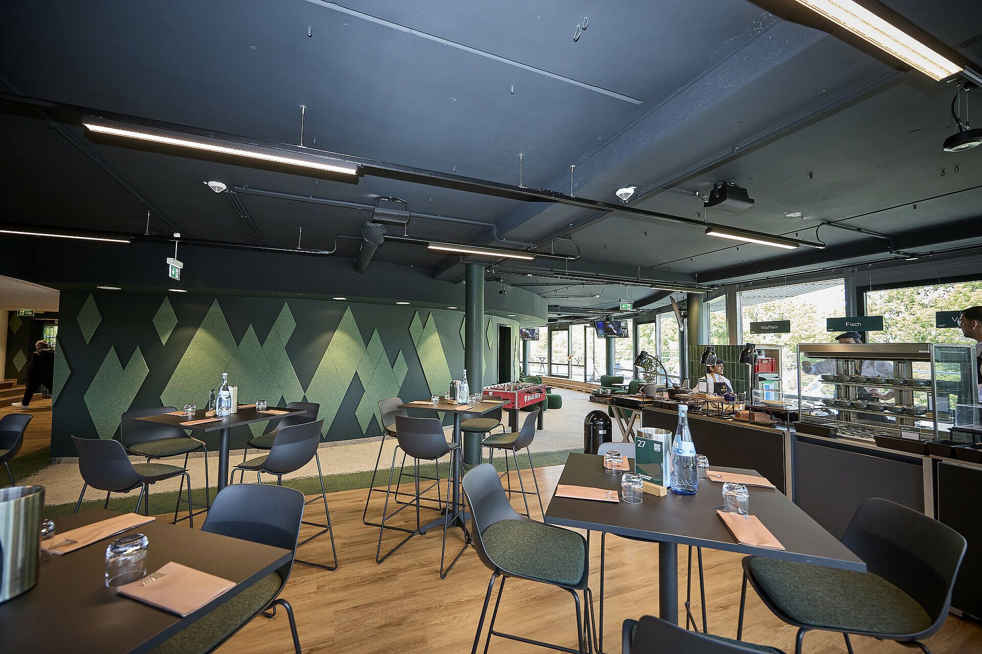 Moderne Cafeteria mit dunkler Decke, geometrischem Wanddesign, Tischen mit Servietten, Wasserflaschen und Stühlen. Große Fenster sorgen für Tageslicht.