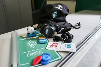 Ein Sensoriktäschchen mit Kopfhörern, Ohrstöpseln, einer Sonnenbrille, Stressbällen, Lernkarten, einem Flyer und einem kleinen Fußball auf einem Tisch.